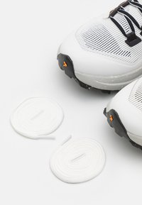 Hvite sportssko med mesh-overdel og svart gummisåle, ledsaget av to løkker av hvite skolisser, med oransje detaljer.