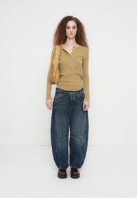 Μπλούζα henley με μακριά μανίκια σε μουσταρδί χρώμα με κουμπιά, συνδυασμένη με φαρδιά μπλε τζιν παντελόνια. Το σύνολο περιλαμβάνει μια υφασμάτινη μπεζ τσάντα ώμου και μαύρα παπούτσια.