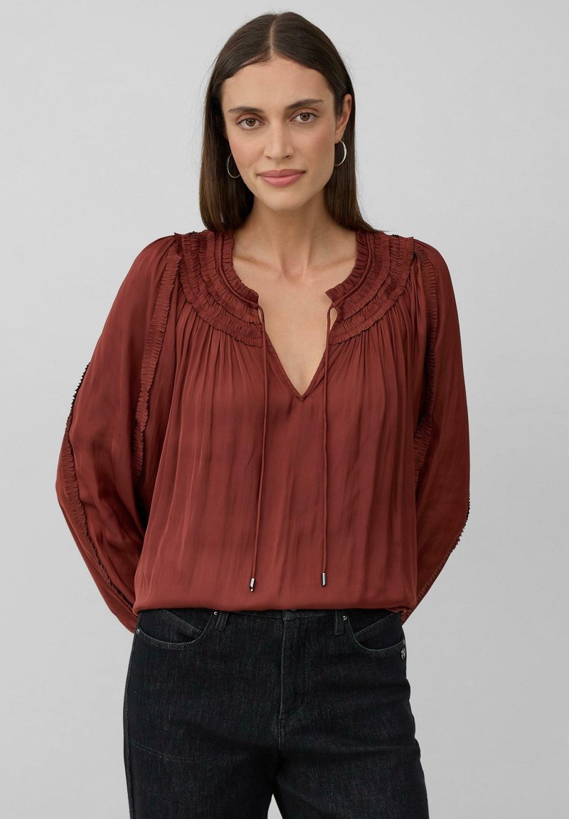 Roestkleurige blouse met een geplooide halslijn, diepe V-hals en verstelbare koorden. Zachte, vloeiende stof met brede mouwen en textuurdetails.