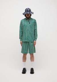 Set format cu un tricou și șorturi în dungi verzi și crem, completat cu o pălărie tip găleată cu model bleumarin, pantofi negri și șosete albe. Casual, cu o croială lejeră.