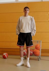 Giocatore di basket con una felpa grigia chiara, una maglietta bianca, pantaloni corti neri con dettagli rossi, calze bianche e sneakers bianche in una palestra.