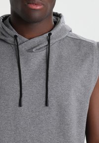 Primer plano de una persona que lleva un hoodie gris sin mangas con cuerdas negras, mostrando los labios y la parte superior del torso contra un fondo simple.