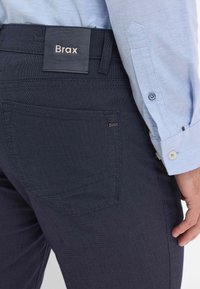 Pantalon texturé bleu marine foncé avec une poche visible et une étiquette en cuir portant la mention "Brax". Une chemise à boutons bleu clair est partiellement visible.