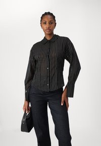 Camicia a righe nere con colletto, maniche lunghe e design aderente, abbinata a jeans scuri e una borsa nera.
