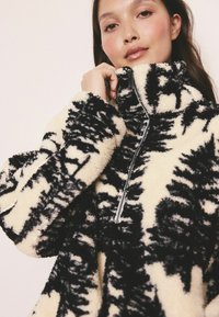 Pullover din fleece, crem cu model abstract negru, având un guler înalt și închidere cu fermoar. Textură moale cu mâneci voluminoase.