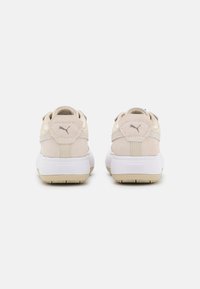 Baskets en suede beige avec des semelles en caoutchouc blanches, de forme arrondie, et des languettes à l'arrière présentant un logo. Texture douce avec des accents contrastés.
