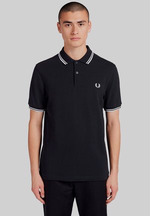 Joven con cabello corto que lleva una camisa polo negra con doble línea blanca en el cuello y las mangas, posando frente a un fondo liso.