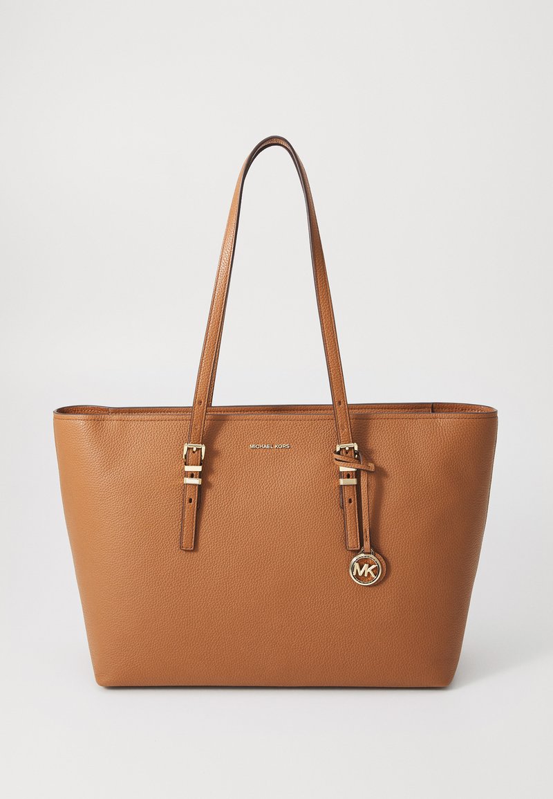Sac fourre-tout en cuir marron avec deux poignées, quincaillerie dorée et une étiquette avec un logo circulaire. Surface texturée avec un design minimaliste.