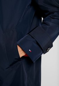 Granatowy wodoodporny płaszcz o gładkiej fakturze, z mankietem zapinanym na guziki oraz małym logo z czerwono-biało-niebieskimi akcentami na rękawie.