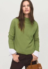 Pull en tricot vert avec un col montant et des poignets contrastants blancs. Porté avec un pantalon sombre, agrémenté d'un sac à main marron et d'accents en anneau.