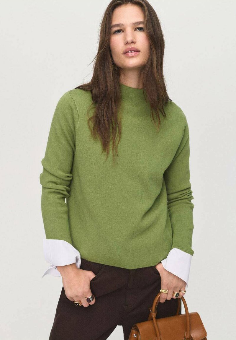 Pull en tricot vert avec un col montant et des poignets contrastants blancs. Porté avec un pantalon sombre, agrémenté d'un sac à main marron et d'accents en anneau.