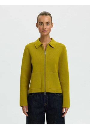 Femme portant une veste jaune moutarde zippée avec poches avant et pantalon bleu foncé, debout devant un fond clair uni.