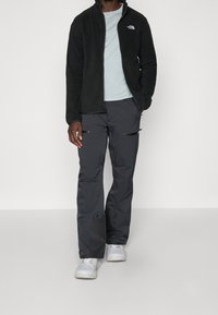 Musta fleece-takki korkealla kauluksella, vaaleansininen t-paita, harmaat housut vetoketjuilla ja vaaleanharmaat lenkkitossut, jotka tarjoavat kerroksellisen ulkoilmeikkauksen.