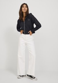 Sweatshirt preto com capuz cortado e fecho, com um bolso frontal e detalhe de logotipo, combinado com jeans brancos de perna larga e ténis pretos.