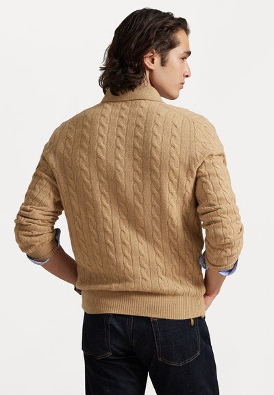 Pull en maille torsadée beige avec un col et des poignets côtelés. Présente un motif texturé, une coupe légèrement ample, et est porté par-dessus une chemise bleue.