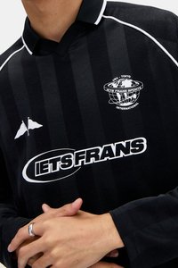 Černý pruhovaný fotbalový dres s bílým límcem a akcenty. Obsahuje logo na hrudi a malý emblém bílých zkřížených vlajek.