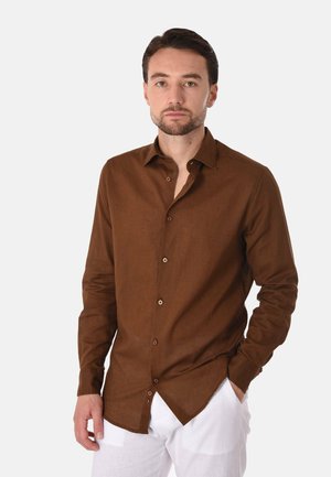 Uomo con capelli corti e scuri e barba, che indossa una camicia marrone abbottonata e pantaloni bianchi, in piedi con una mano in tasca davanti a uno sfondo bianco.