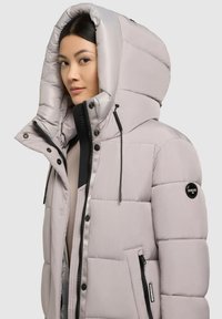 Giacca a vento grigio chiaro con un grande cappuccio, chiusura frontale con zip e dettagli neri. Presenta un design trapuntato e tasche laterali. Materiale impermeabile.