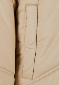 Beige polstret jakke med sydd mønster, en rektangulær lomme og glatt tekstur. Uthevede sømmer forbedrer strukturen.