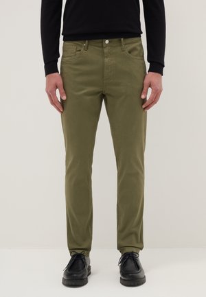 Man draagt olijfgroene slim-fit broek en zwarte veterschoenen, staand tegen een effen lichte achtergrond.