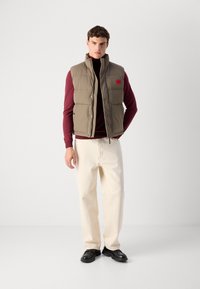 Man wearing beige padded vest over burgundy sweater, off-white pants, and black dress shoes standing against a plain light background.