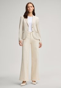 Jasno beżowa marynarka o dopasowanym kroju z dwoma guzikami, zestawiona z kremowymi spodniami typu wide-leg i jasnymi obcasami, na tle neutralnym.