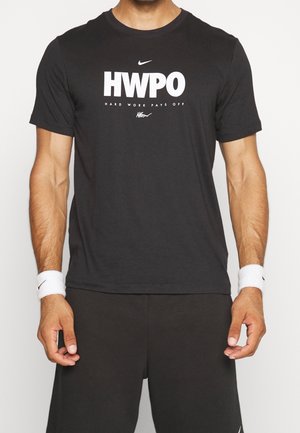 Mann trägt ein schwarzes Nike-T-Shirt mit dem Schriftzug "HWPO Hard Work Pays Off" und weißen Handgelenkbändern an beiden Handgelenken, steht vor einem weißen Hintergrund.