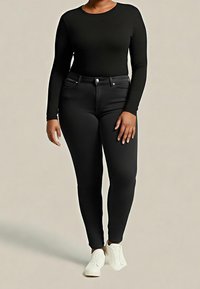 Personne portant un haut noir à manches longues, un jean noir ajusté et des baskets blanches, debout devant un fond clair uni.