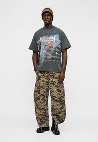 Mees, kes kannab musta mütsi, tuhmunud graafilise kujundusega T-särki, kamuflaažkujulisi püksid ja musti läikivaid saapaid, seisab valge tausta ees.