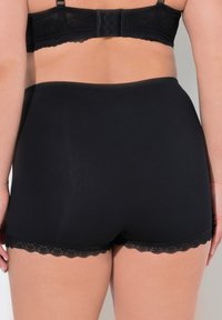 Mutandine nere a vita alta con una texture liscia e dettagli in pizzo lungo le aperture delle gambe, viste dal retro.