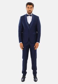 Tuxedo navy con rever a scialle a motivo, camicia bianca e cravatta a farfalla nera. Pantaloni attillati e scarpe nere lucide completano l'insieme.