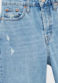 Jean en denim bleu clair avec une texture usée, dotés d'une petite poche avant, de rivets en cuivre et de déchirures subtiles au niveau des cuisses.