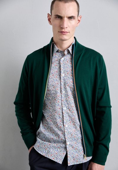 Grønn strikket cardigan med glidelås, kombinert med en blomstret skjorte med knapper som har små røde og blå blomster på hvit bakgrunn.