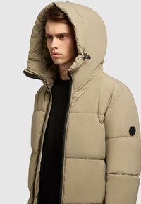 Beige puffjakke med en stor hætte, lynlåslukning og sorte detaljer. Har et quiltet design og et rundt sort logo på venstre ærme.