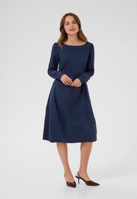 Jūros laivyno ilgomis rankovėmis suknelė su kvadratine iškirpte, surinktomis rankovėmis ir plačiu sijonu. Dėvima su burgundiškai raudonais smailiais bateliais.