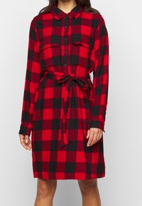 Vestito camicia a scacchi rossi e neri realizzato in tessuto morbido, dotato di maniche lunghe, chiusura con bottoni sul davanti, due tasche sul petto e vita regolabile con nastro.