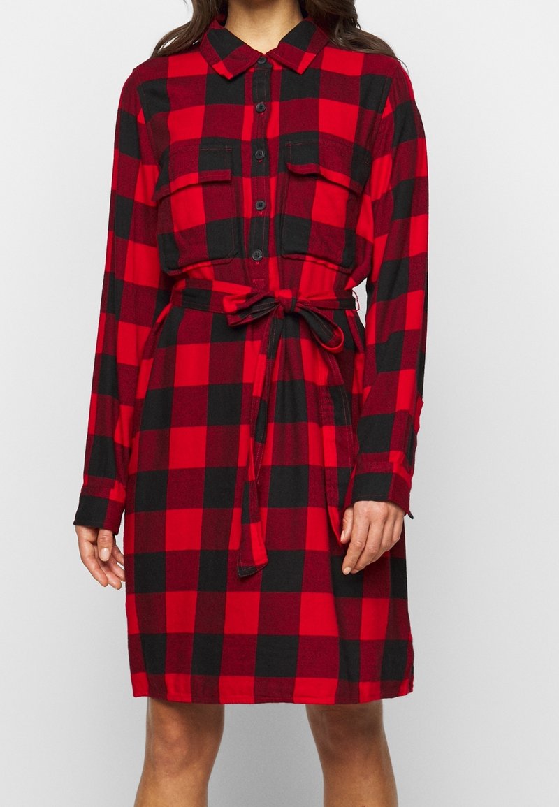 Vestito camicia a scacchi rossi e neri realizzato in tessuto morbido, dotato di maniche lunghe, chiusura con bottoni sul davanti, due tasche sul petto e vita regolabile con nastro.