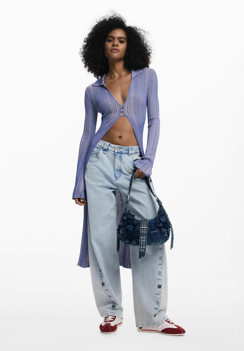 Világos lila bordázott zipzáros felső, magas gallérral, nyitott elülső résszel. Laza kék farmernadrág gombos részletekkel. Tengerészkék textúrájú kézitáska, piros-fehér sportcipő.