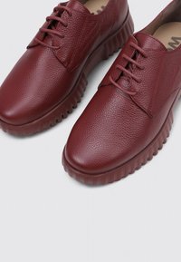 Un par de zapatos de cuero texturizado de color rojo oscuro, con cordones y suelas de goma gruesas y acanaladas, sobre un fondo claro y liso.