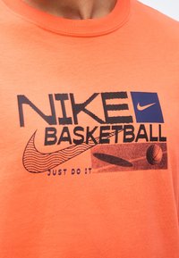 Πορτοκαλί μπλούζα με το κείμενο «Nike Basketball», το λογότυπο swoosh της Nike με κύματα, μια γραφική παράσταση καλαθοσφαίρισης και το σύνθημα «Just Do It» σε μαύρο και μπλε.