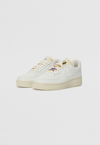 Par de zapatillas blancas de corte bajo con suelas beige y adornos de charms de encaje en verde, azul y oro sobre un fondo sencillo.