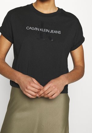 Osoba nosząca czarny krótki top z podwiniętymi rękawami i logo "Calvin Klein Jeans", w połączeniu z oliwkową spódnicą, ręce złożone z przodu.