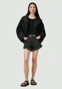 Svart oversized jacka, svart topp, svarta denimshorts med slitna kanter och fransar, svarta sandaler med spännen, modell stående mot en neutral bakgrund.