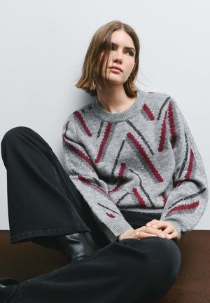 Sivý pletený sveter s výraznými cik-cak vzormi v bordovej a čiernej farbe. Rebrovaný výstrih a manžety. Kombinovaný s čiernymi nohavicami s vysokým pásom.