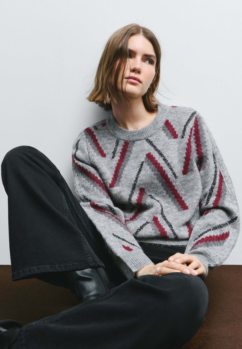 Gray knit sweater featuring bold, zigzag patterns in burgundy and black. Ribbed neckline and cuffs. Paired with high-waisted black pants.