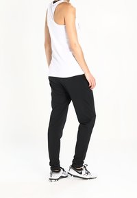 Débardeur blanc sans manches, pantalon noir fuselé et chaussures de sport noires avec détails blancs, vus de dos. Texture du tissu lisse.