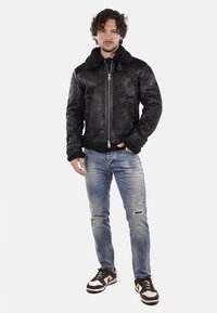 Giacca di pelle nera con colletto in pelliccia, chiusura a zip anteriore e due tasche laterali. Indossata sopra jeans chiari strappati blu e sneakers marroni.