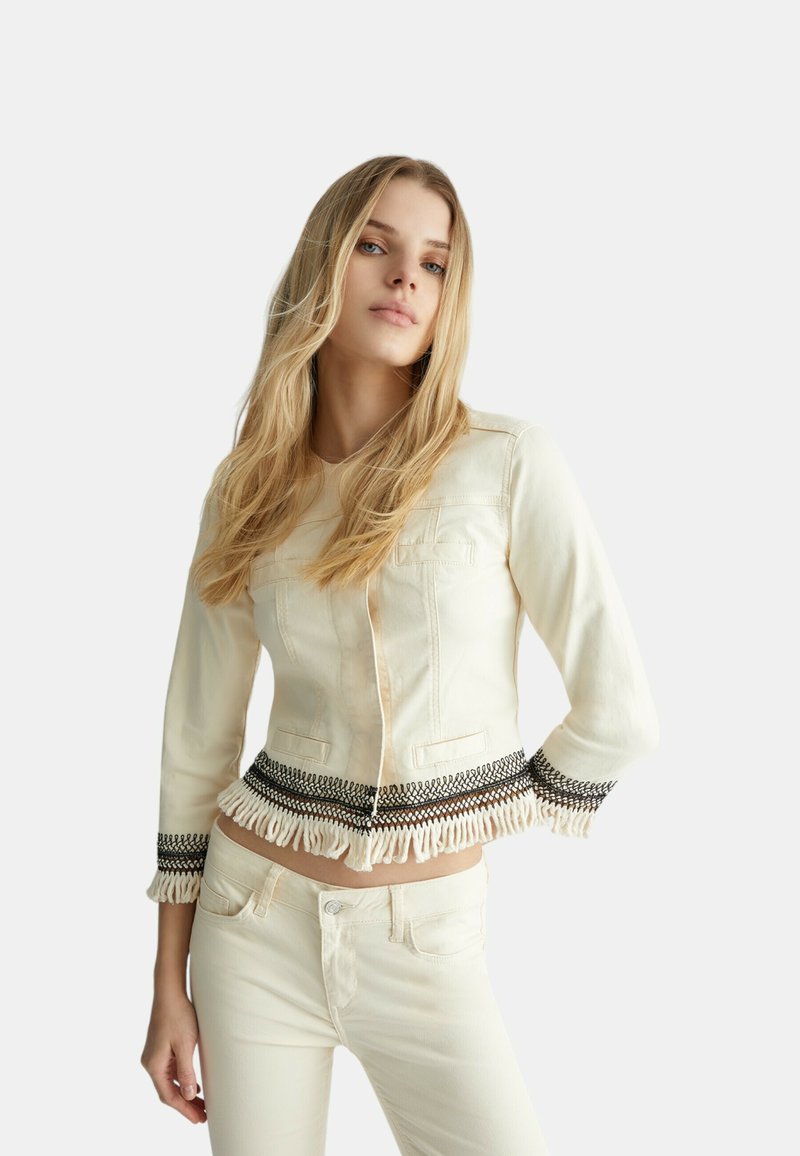 Veste en denim crème au design ajusté, avec des accents brodés noirs complexes et un ourlet frangé. Manches longues, poches avant.