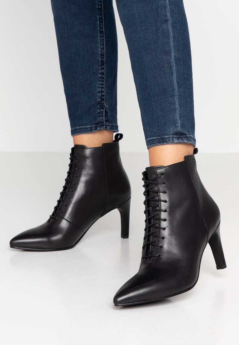 Botas de tobillo de cuero negro con punta afilada, diseño con cordones y un tacón stiletto de 7 cm. Textura suave con un brillo sutil.