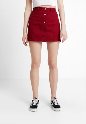 Kvinde iført en mørkerød mininederdel med højtaljet, knaplukning foran, stylet med sorte og hvide sneakers og en hvid cropped top.